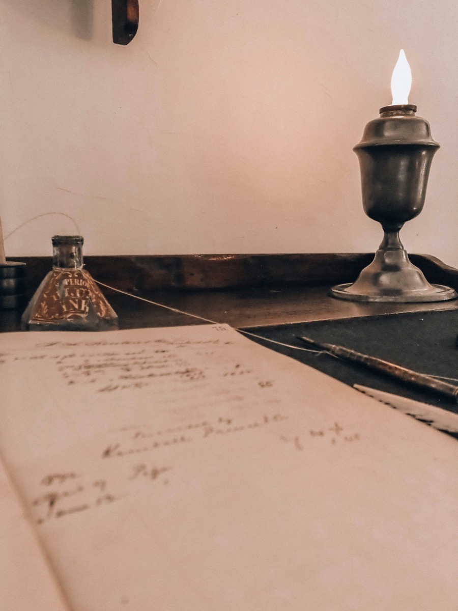 Wooden desk with an open ledger book, a fountain pen and a lamp casting soft light over handwritten pages.